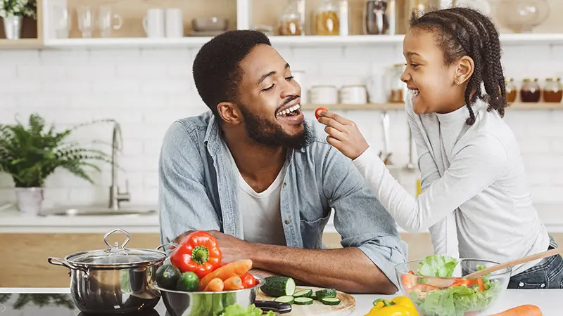 girl giving her dad cherry tomato while cooking